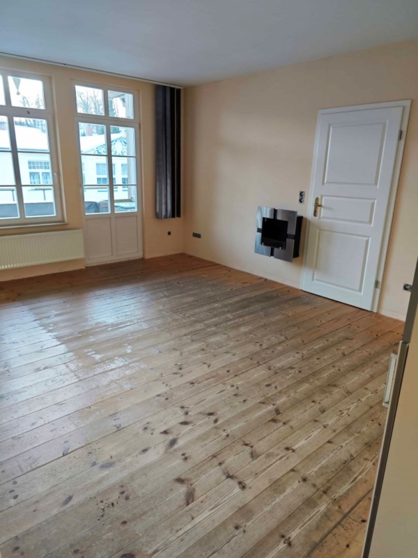 Zimmer vor der Renovierung mit Holzboden, großen Fenstern und beigen Wänden